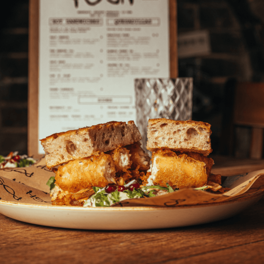 Brown, wooden table with a white plate on top of it, that has a brown bread sandwich on top of brown greaseproof paper